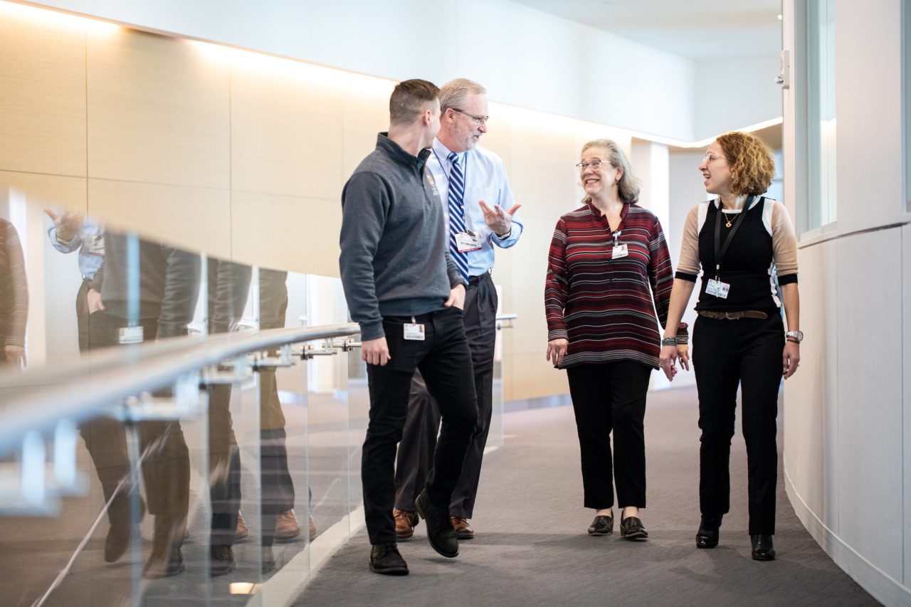 A group of people wearing IDs on clips or lanyards walk down a hallway