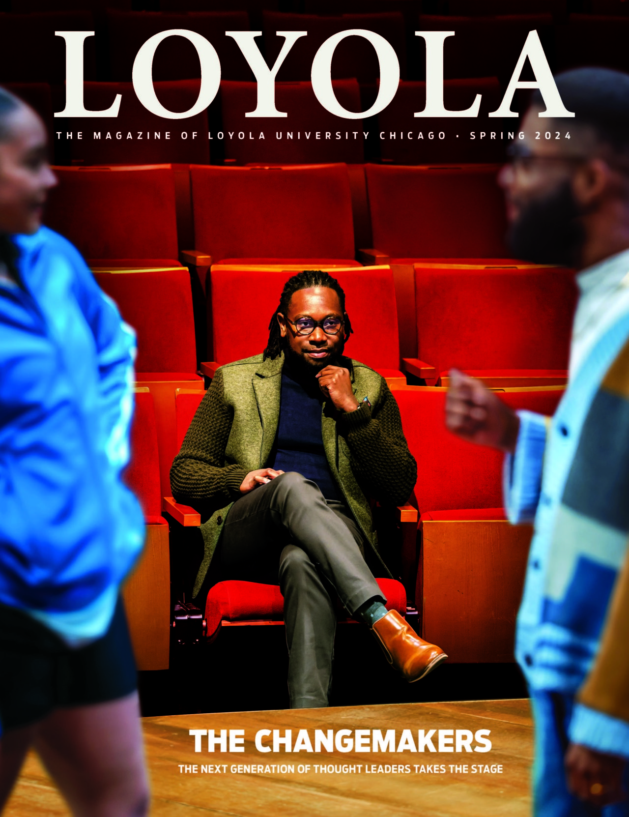 A man wearing a suit sits in red theater seats and rests his head on his hand as he watches students in front of him rehearse a play
