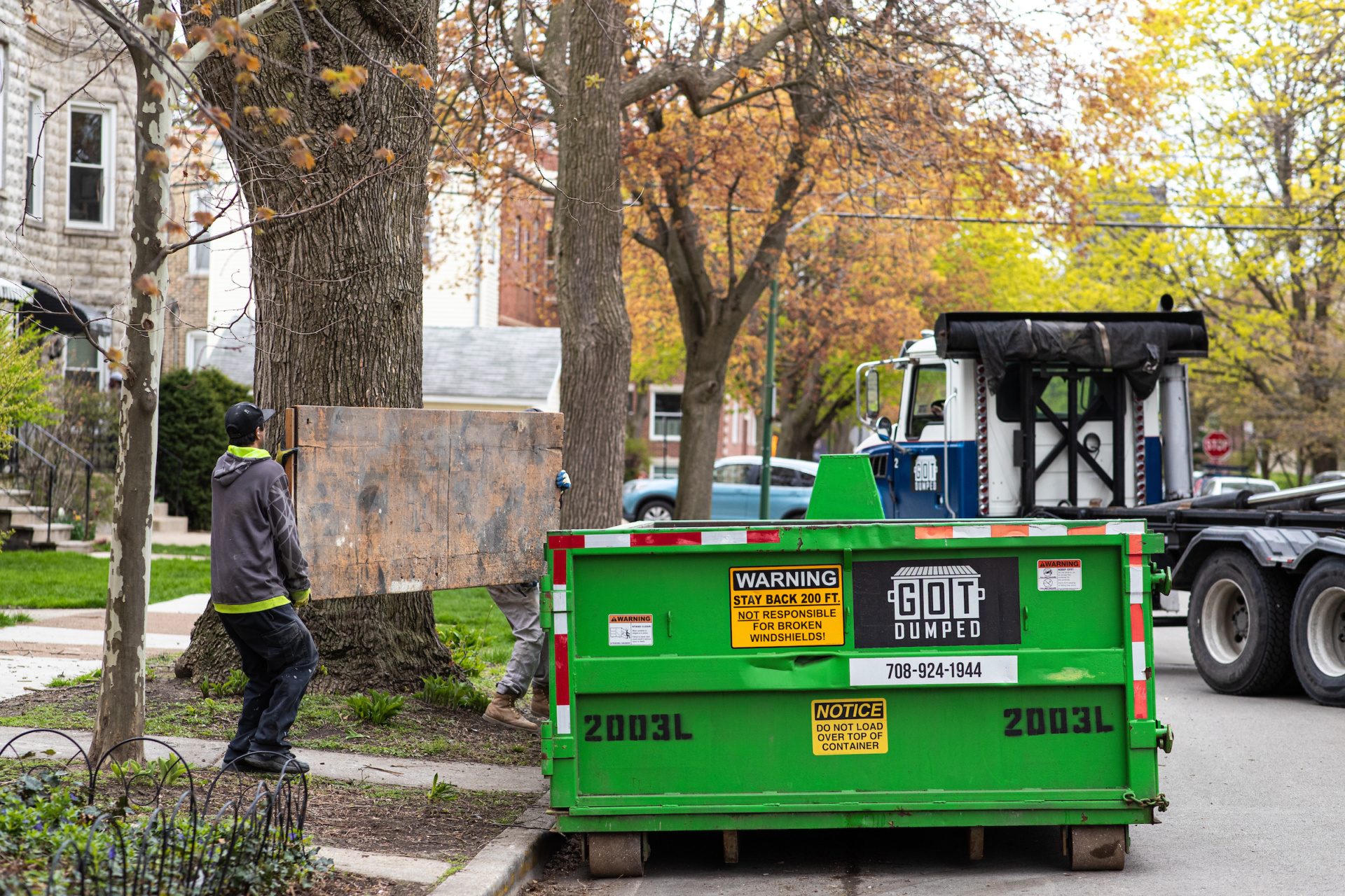 Two men lift a large piece of wood into a large green dumpster on a curb
