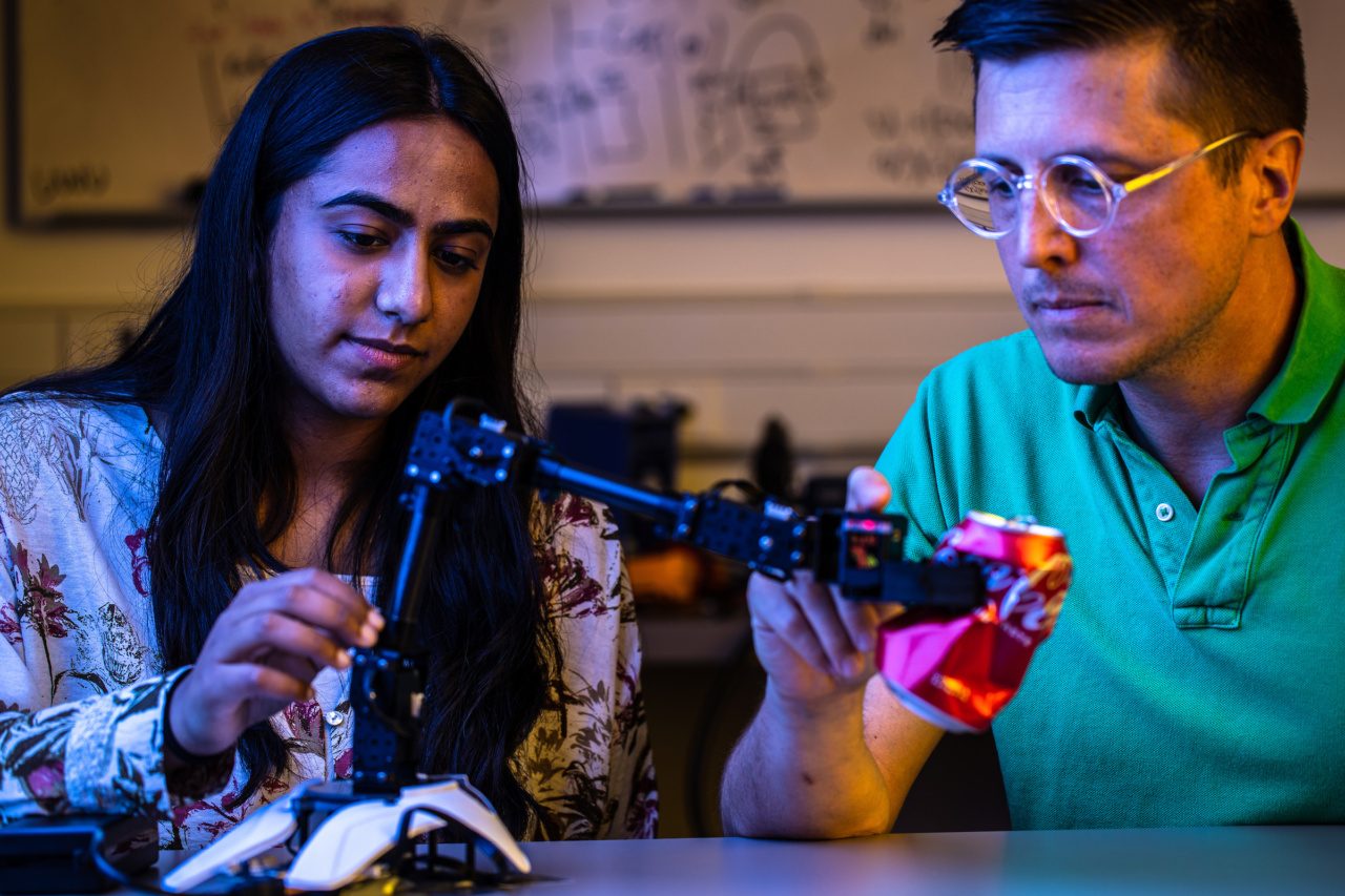 A man and a woman look at a robotic arm crushing a can of Coca-Cola