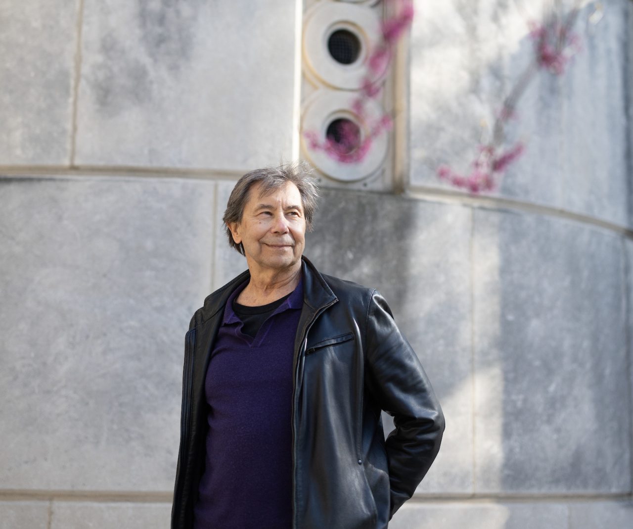 A man wearing a leather jacket and navy T-shirt stands with his hand on his hip in front of a stone wall on the Loyola University Chicago campus with blooming tree branches in the foreground