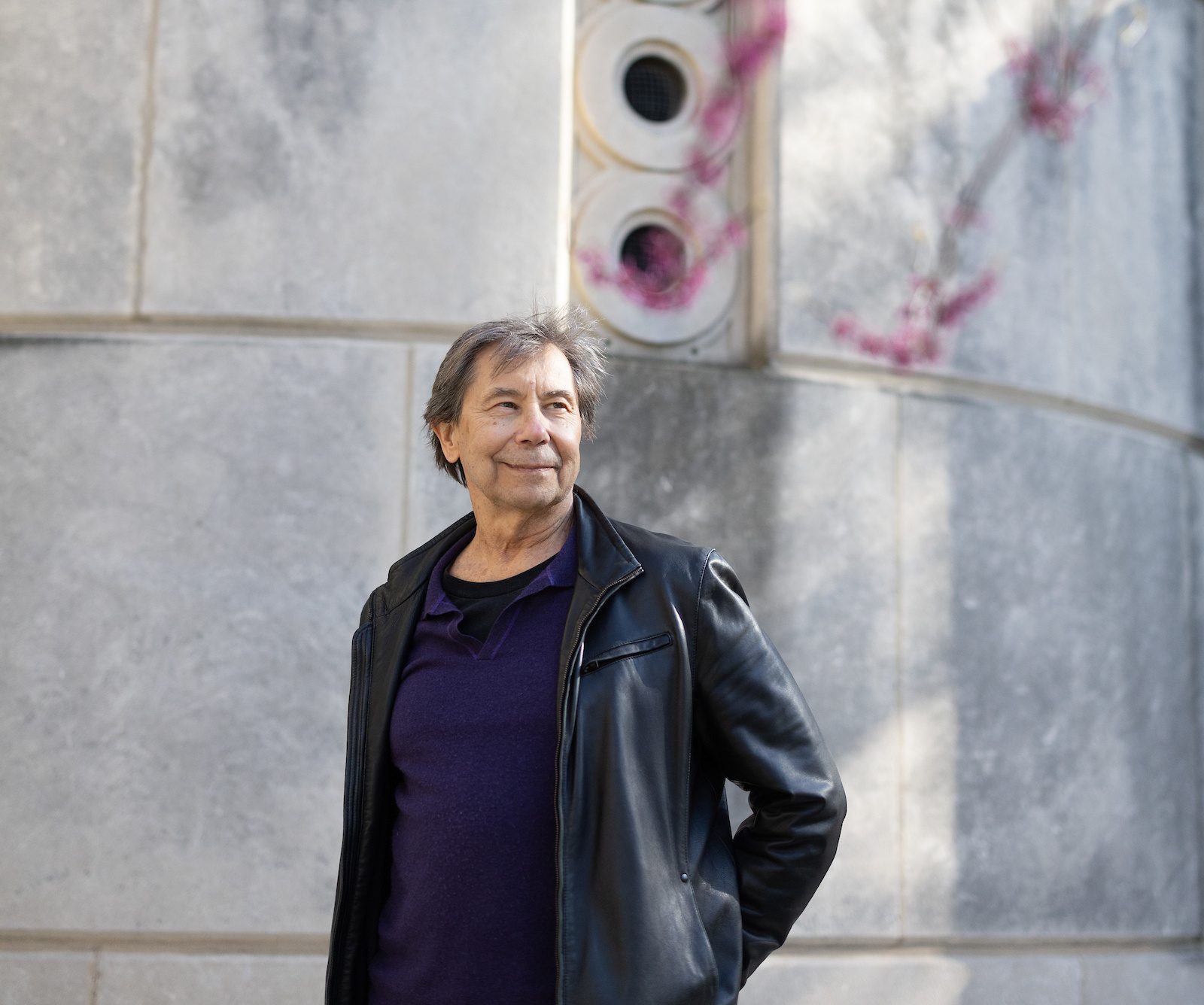 A man wearing a leather jacket and navy T-shirt stands with his hand on his hip in front of a stone wall on the Loyola University Chicago campus with blooming tree branches in the foreground
