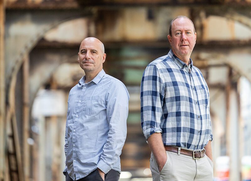 Two man stand back to back with their hands in their pockets under elevated train tracks