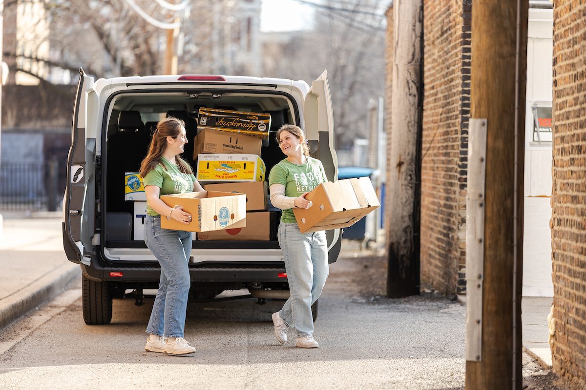 Two Loyola University Chicago students carry cardboard boxes into a building from the back of a white van