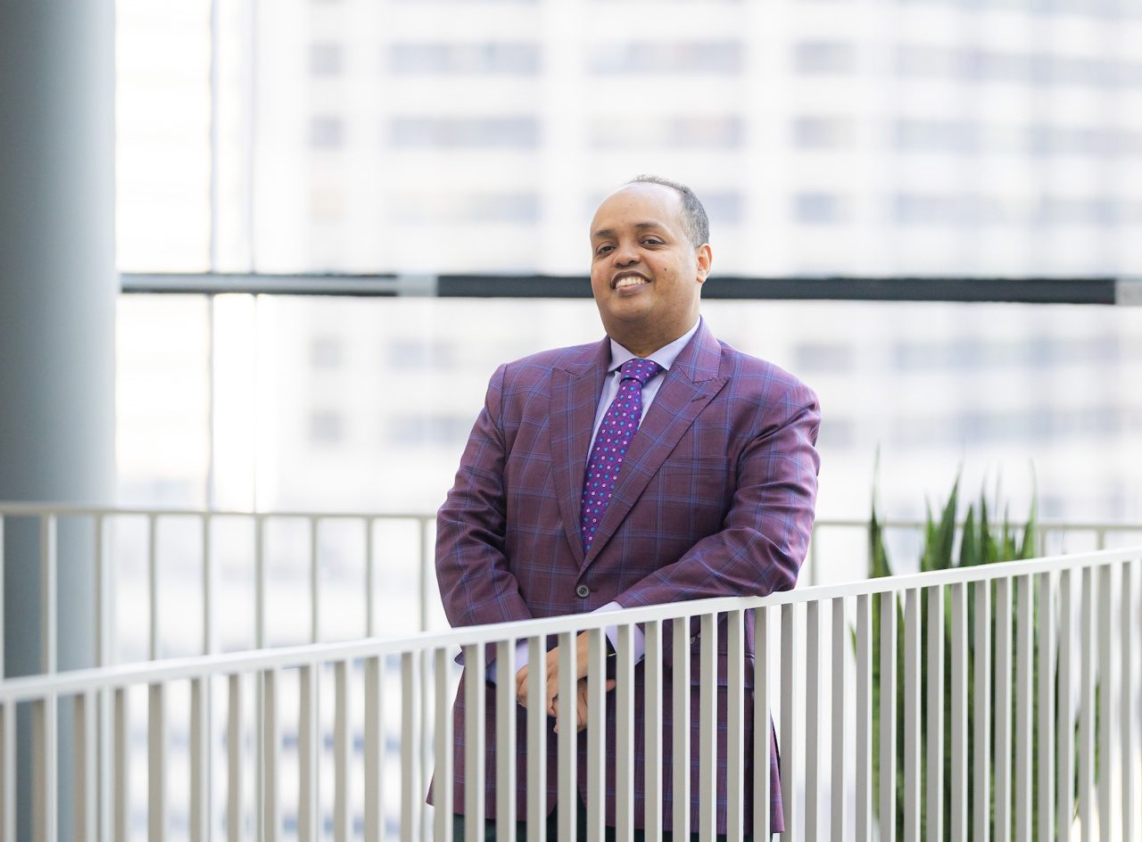 A man wearing a purple suit stands near a white railing in a glass-walled building on the Loyola University Chicago campus