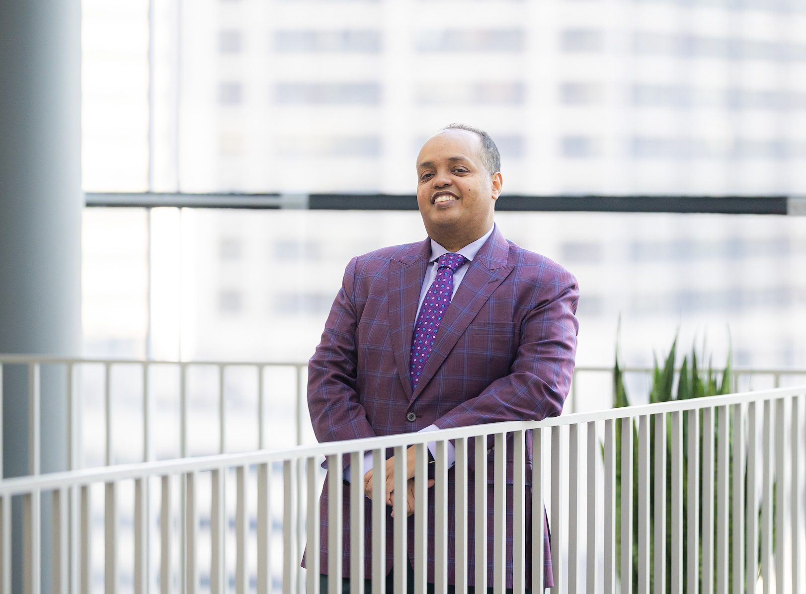 A man wearing a purple suit stands near a white railing in a glass-walled building on the Loyola University Chicago campus