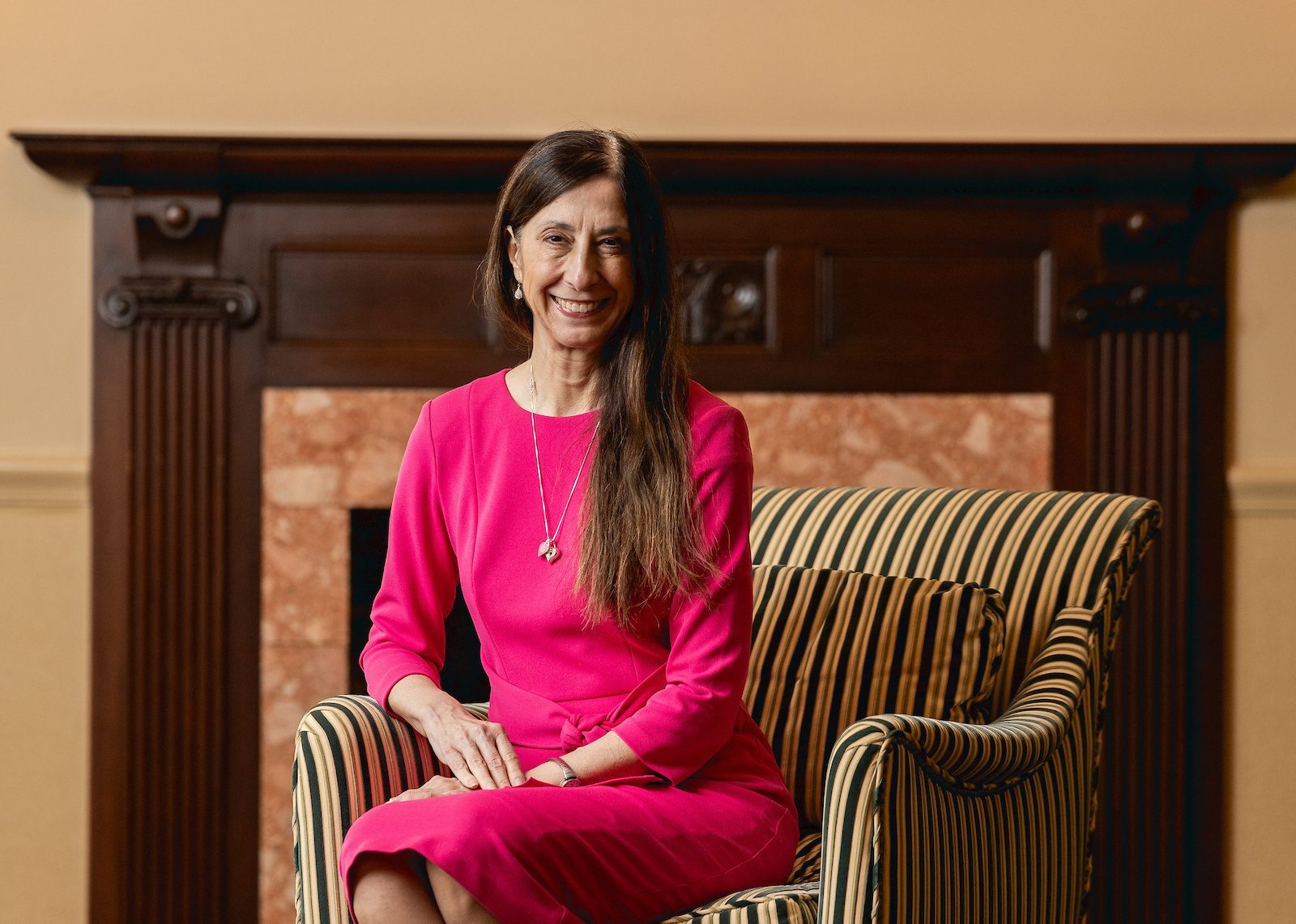 A woman in a hot pink dress smiles as she sits in a striped armchair