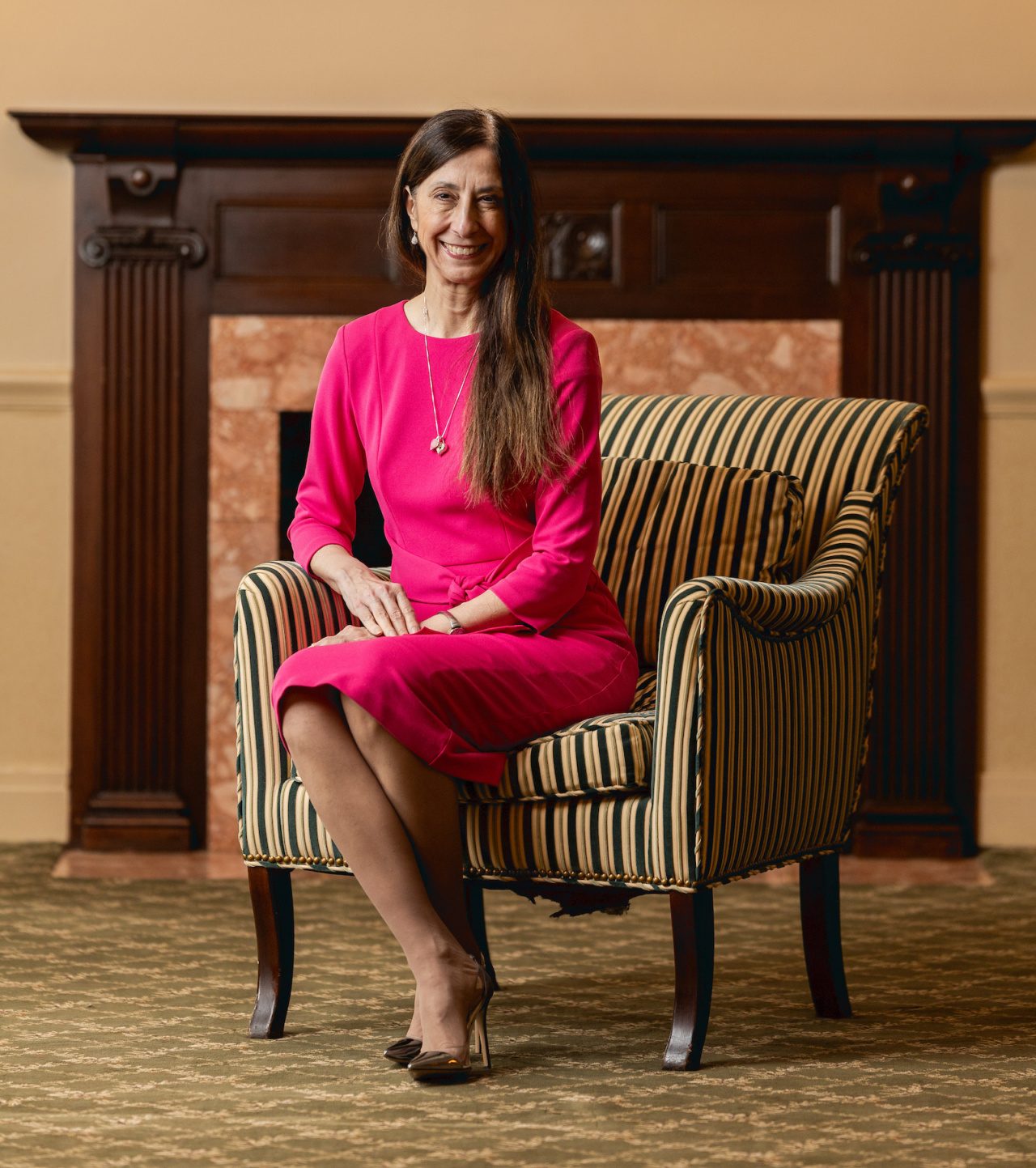 A woman wearing a hot pink dress sits in a striped armchair in front of a fireplace